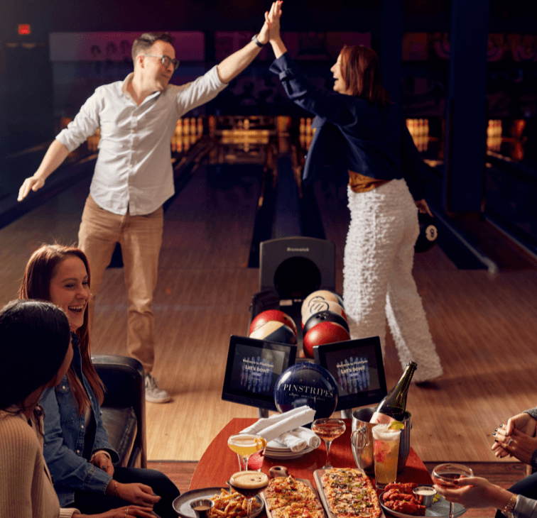 Friends at a Pinstripes bowling lane, two high-fiving in the background while others laugh over food and drinks in the foreground