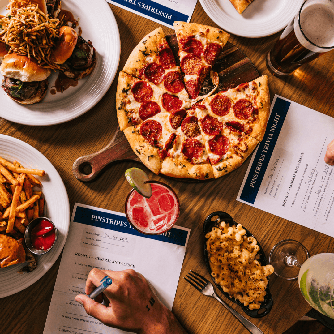 Friends fill out trivia cards beside pizza, cocktails, mac and cheese and Tenderloin Sliders.