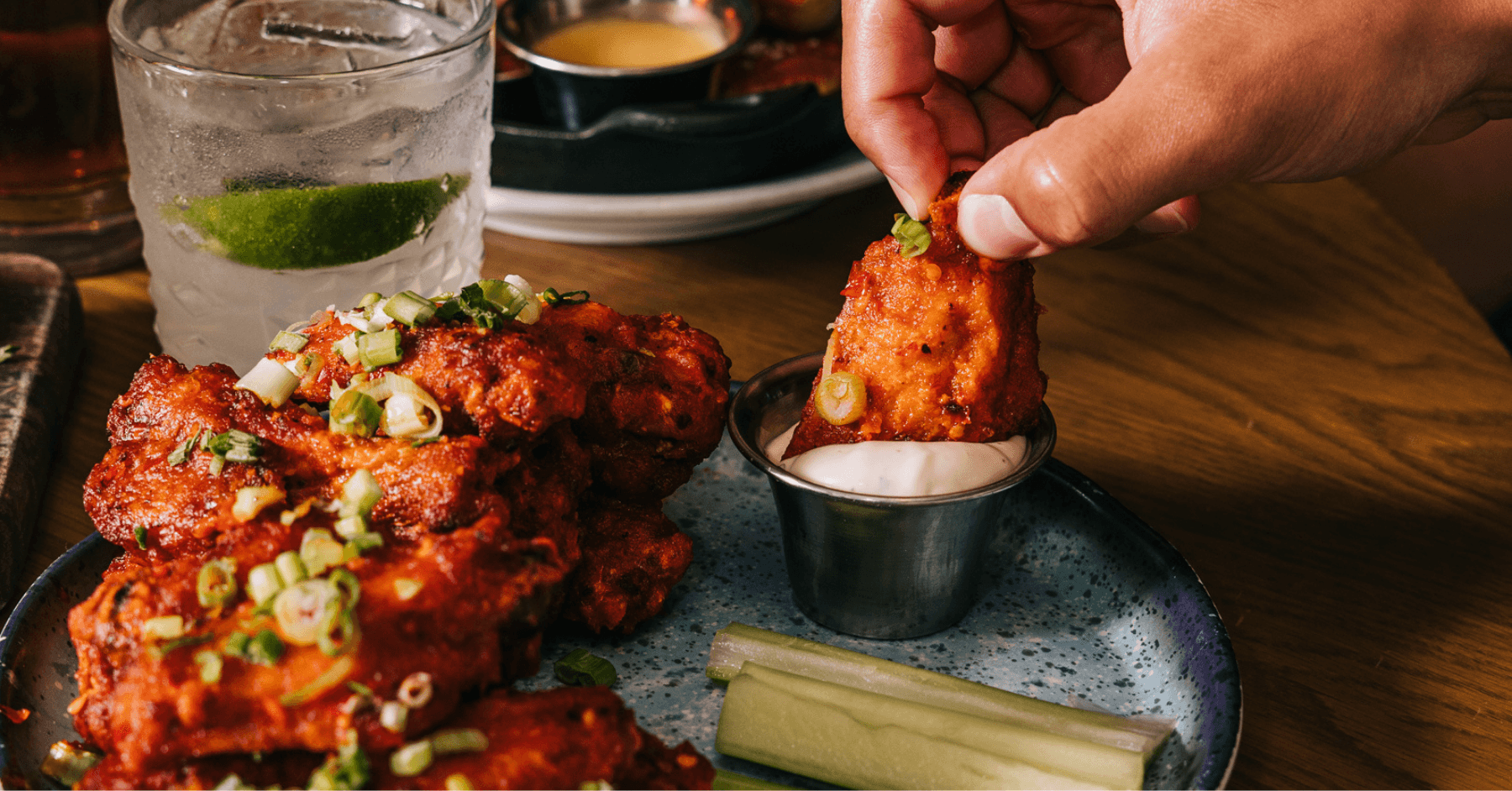 Hand dipping a Pinstripes Signature Wing into ranch with a cocktail in the background.