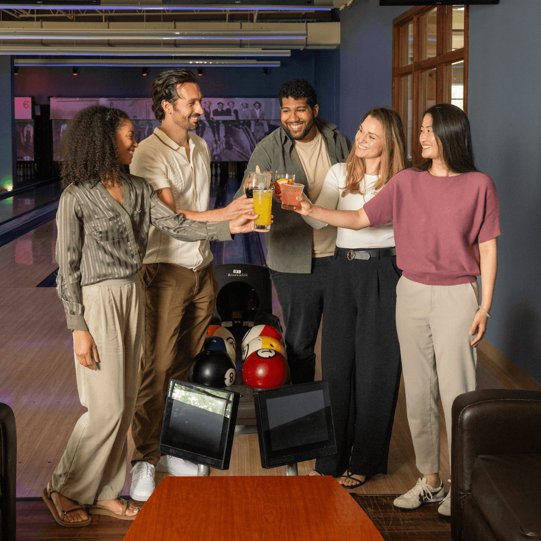 Five friends raising drinks together over the bowling lanes at Pinstripes.