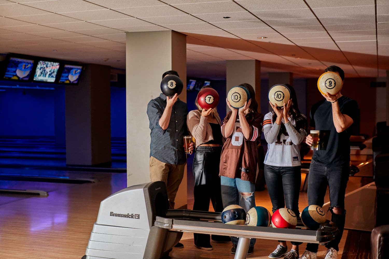 Five friends on Pinstripes bowling lanes being silly, each holding a bowling ball in front of their face, highlighting the fun, social side of bowling with friends.