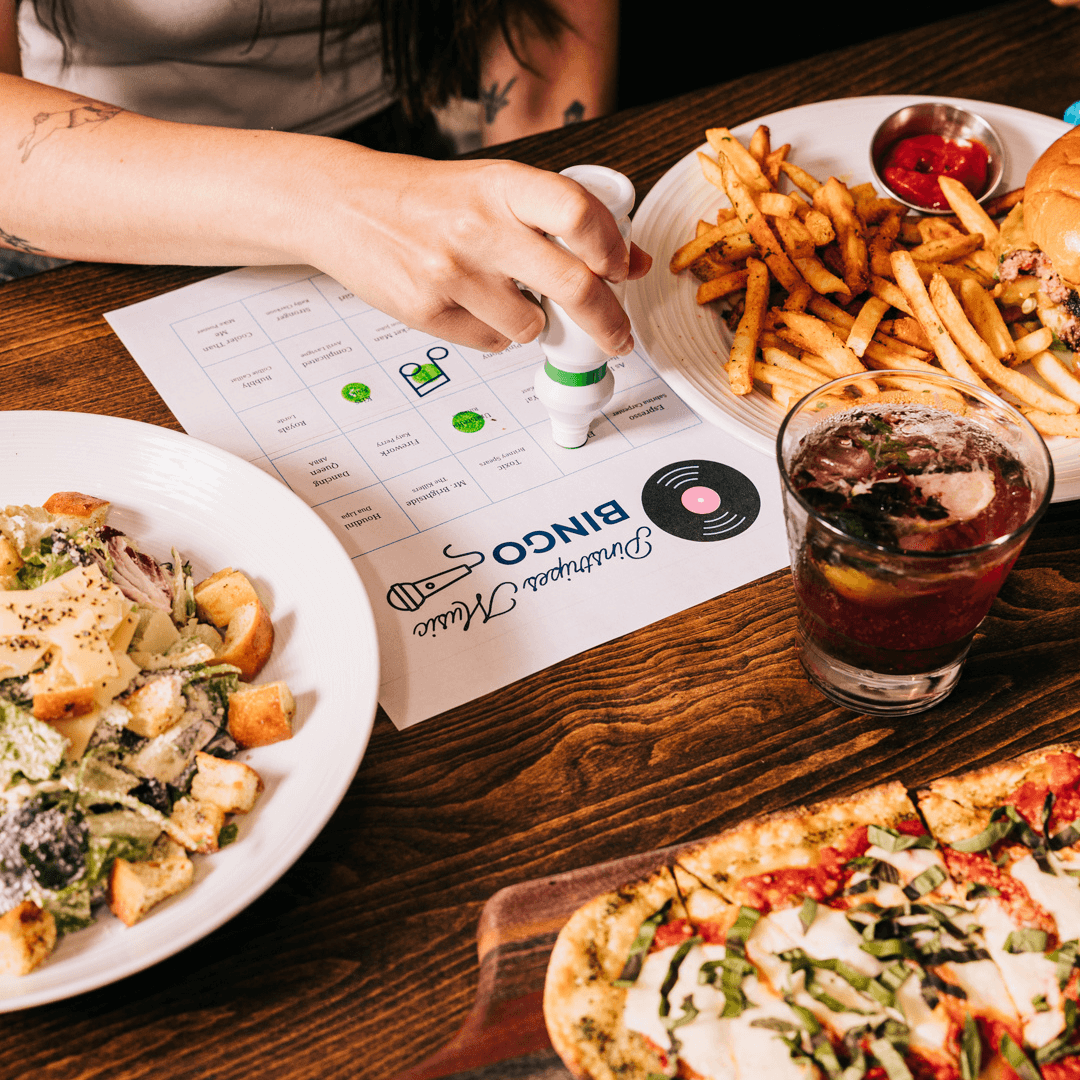 Music bingo card on a table surrounded by Caesar salad, pizza, a cocktail and other dishes.