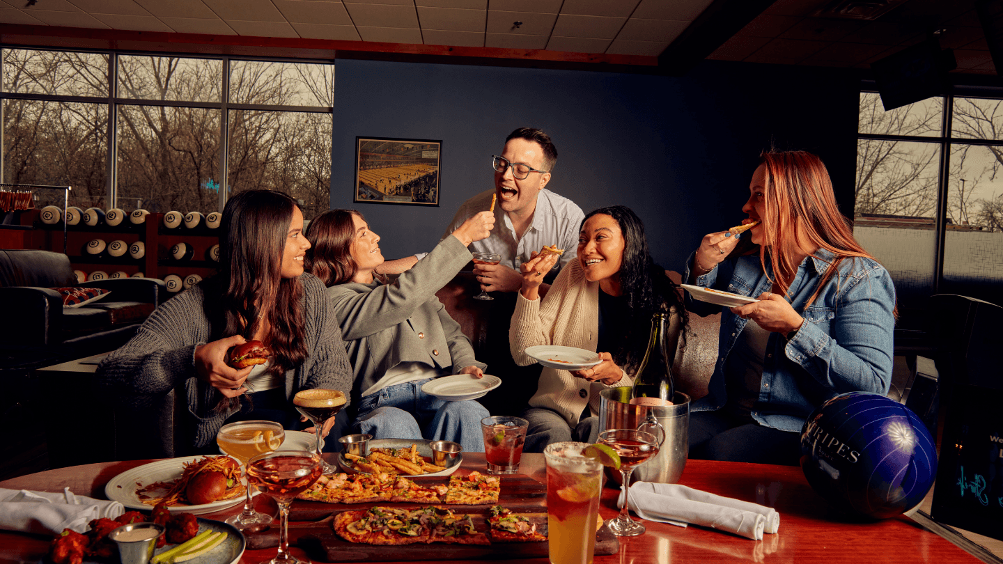Friends sitting on couch at Pinstripes bowling lane laughing and playfully feeding each other with food spread