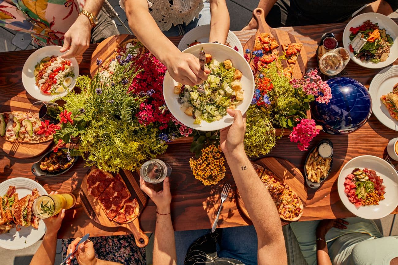 Large shared table at Pinstripes Bistro filled with flatbreads, salads, and shareable dishes, with a group of friends passing plates and two friends in the center passing a Caesar salad.