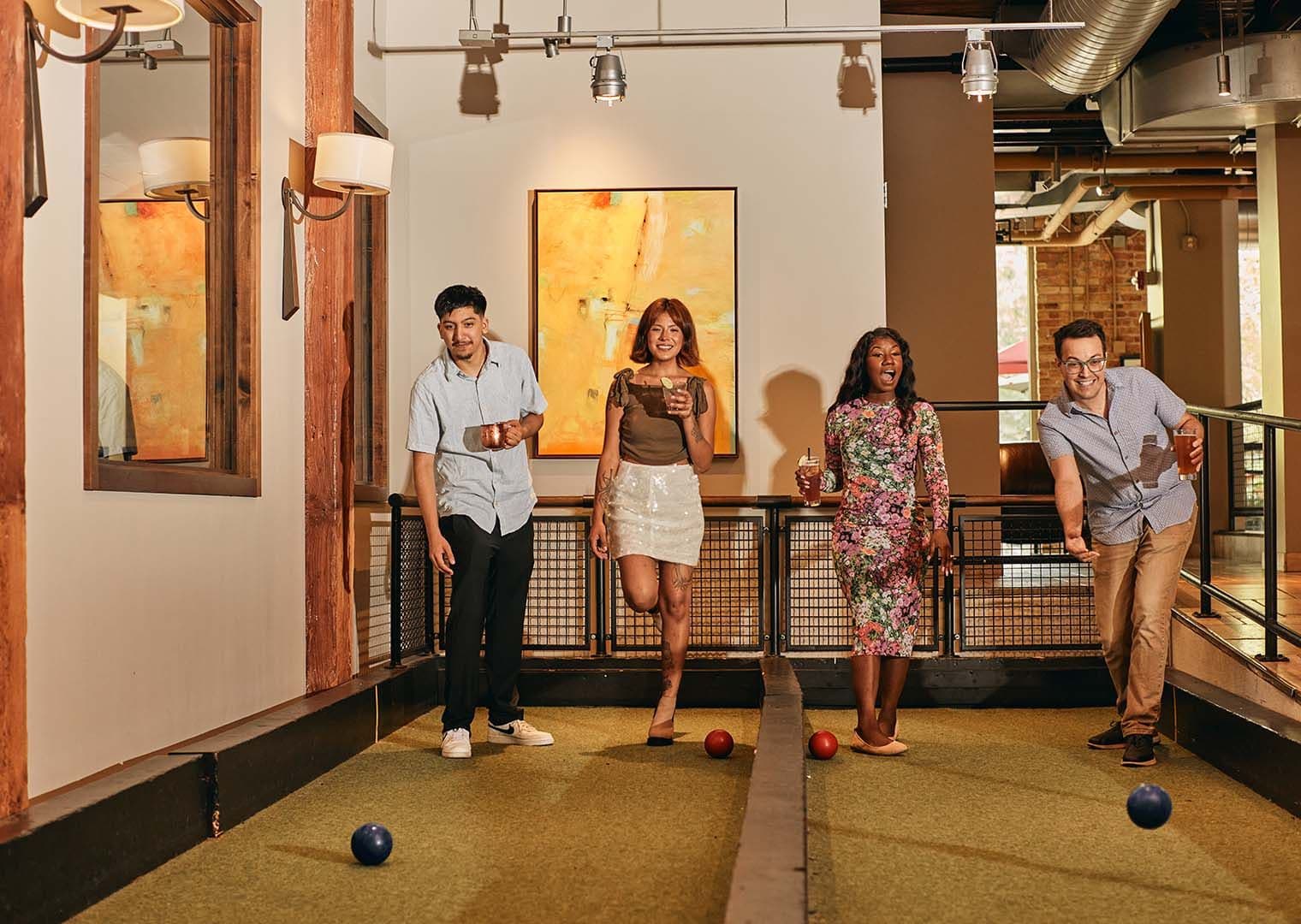 Four friends playing bocce together on a bocce court, each holding handcrafted cocktails, highlighting social, relaxed outdoor‑style game time.