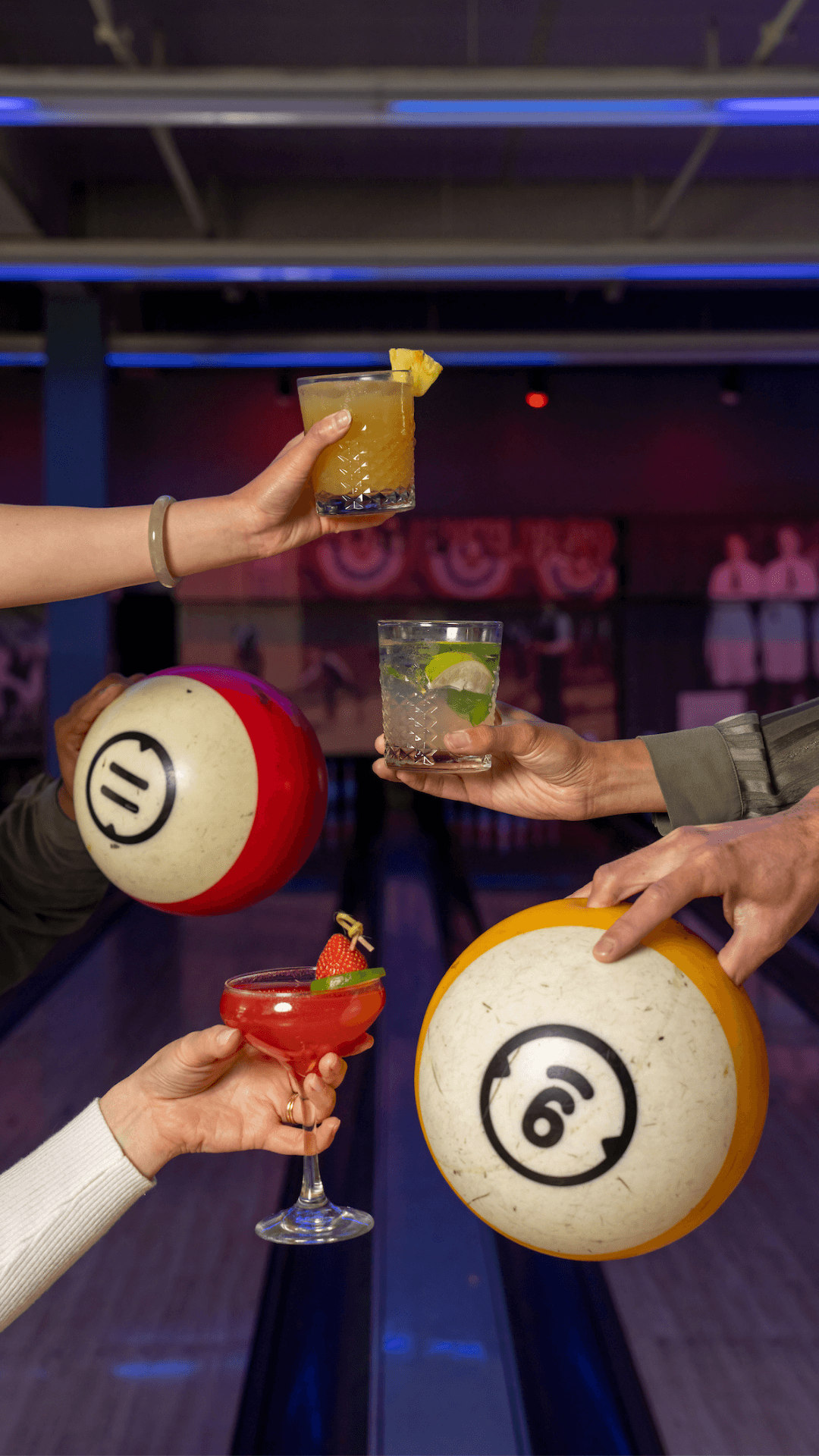 Two hands holding drinks and two holding bowling balls together above the bowling lanes.