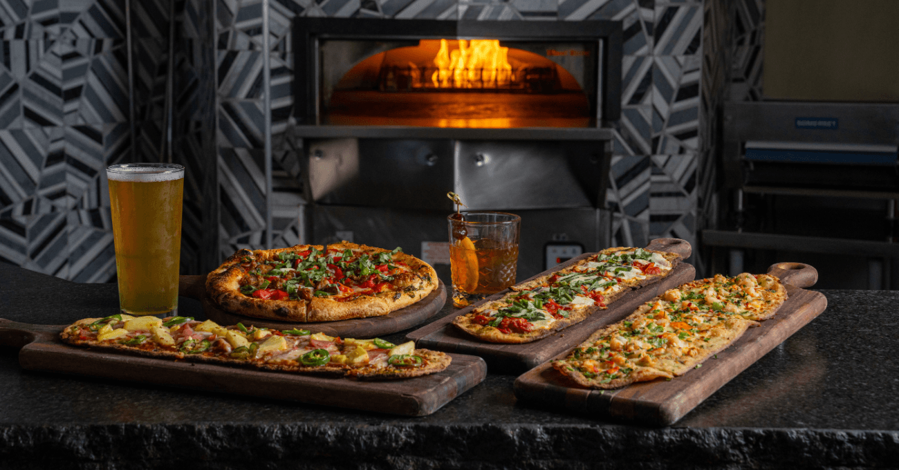 Sausage and Peppers Pizza, Hawaiian, Margherita and Lobster flatbreads with beers in front of a wood-fired oven.