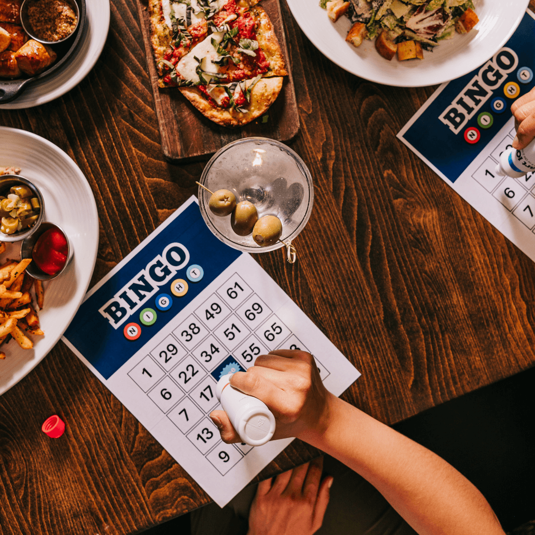 Guest marks a bingo card with a dauber beside flatbreads, pretzel bites and a Dirt Martini.